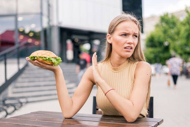 美丽年轻悲伤的女人拿着汉堡包坐在户外吃快餐不满意微笑街道午餐
