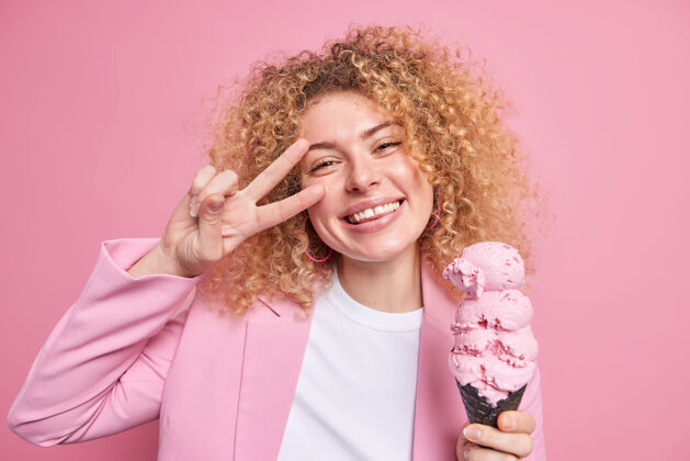 高兴积极漂亮的女人卷发吃巨大的冰淇淋在圆锥形华夫饼使和平姿态微笑广泛有乐趣在夏日穿着时髦的衣服隔离在粉红色的墙壁脸微笑女性