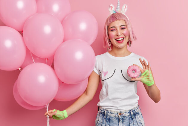 心情高兴的粉红色头发的女人拿着一堆气球美味的甜甜圈微笑愉快地享受在聚会上的空闲时间穿着休闲t恤牛仔裤运动手套微笑穿节日