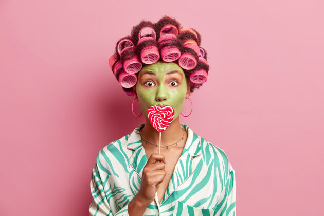 心情震惊的非裔美国女人的室内镜头用卷发器做完美的发型用棒棒糖盖住嘴做绿色滋养面膜减少隔离在粉色墙壁上的细纹室内时尚封面