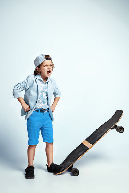 童年在白色的工作室墙上 穿着休闲服的漂亮男孩在滑板上男孩站活跃