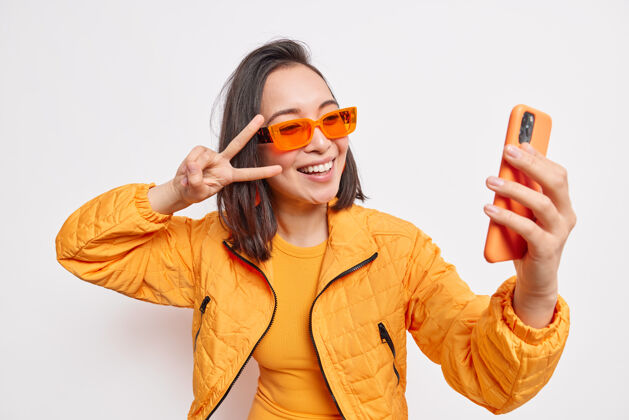 夹克美丽时尚的亚洲女人在手机上自拍让v标志微笑有正面的脸戴着橙色太阳镜和夹克隔着白墙现代的生活方式姿势快乐欢呼