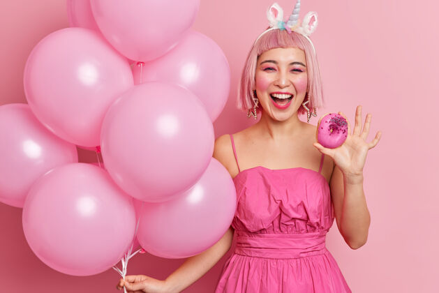 乐观快乐的亚洲年轻女子的照片有粉红色的头发穿着喜庆的裙子拿着美味的甜甜圈和一堆充气气球享受派对时间女性少女束