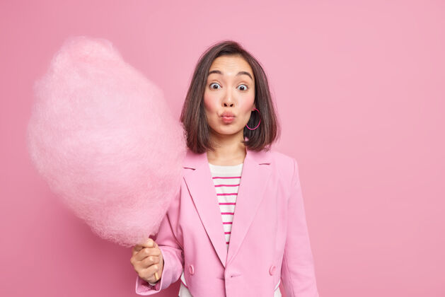 模特人与休闲的概念可爱的深色头发的亚洲女人保持嘴唇合拢举行美味的糖果牙线棒上穿着时尚的衣服隔离在粉红色的墙壁上蓬松糖亚洲