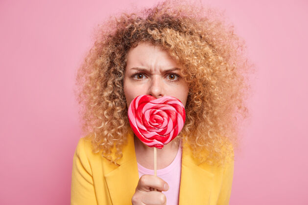 卷曲爱吃甜食的概念严肃的卷发年轻的欧洲女人用美味的棒棒糖捂着嘴 用担心的表情集中不高兴的姿势对着粉色的墙壁时尚饮食紧张