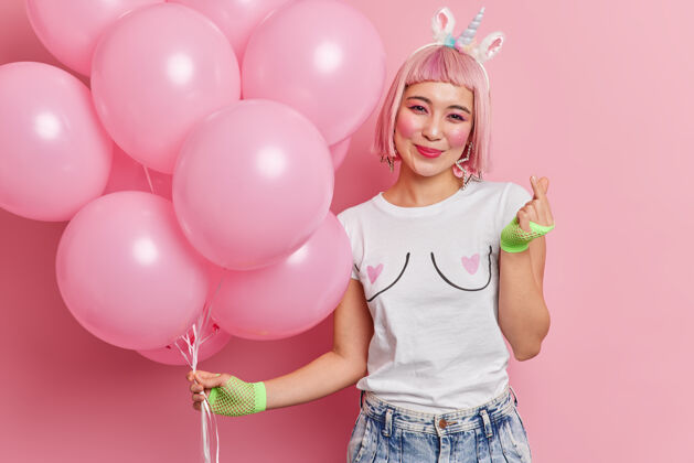 欢呼快乐的粉红色头发的女人的肖像与东方外观手势迷你标志手势姿势与氦气球享受节日穿着时髦的衣服快乐美丽微笑