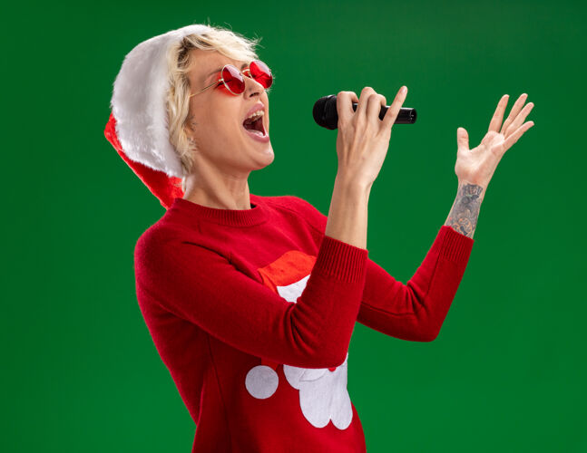 穿年轻的金发女郎戴着圣诞帽和圣诞老人的圣诞毛衣戴着眼镜站在侧视图拿着麦克风手在空中唱歌闭着眼睛隔离在绿色的墙上女人麦克风保持