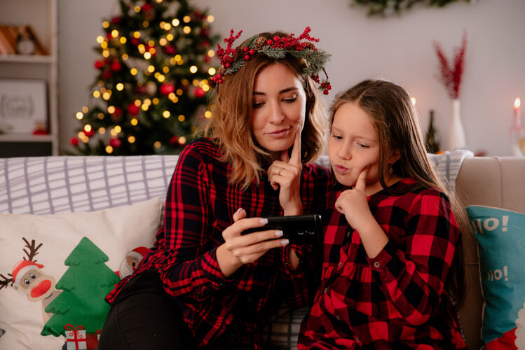 房子体贴的母亲和女儿坐在沙发上看着电话 在家里享受圣诞节时光享受电话沙发