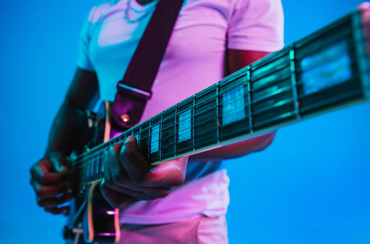 男性年轻的非洲裔美国音乐家在霓虹灯下像摇滚明星一样在蓝色的工作室背景上弹吉他音乐的概念 爱好快乐的家伙即兴创作复古的彩色肖像音乐会酒廊灯光
