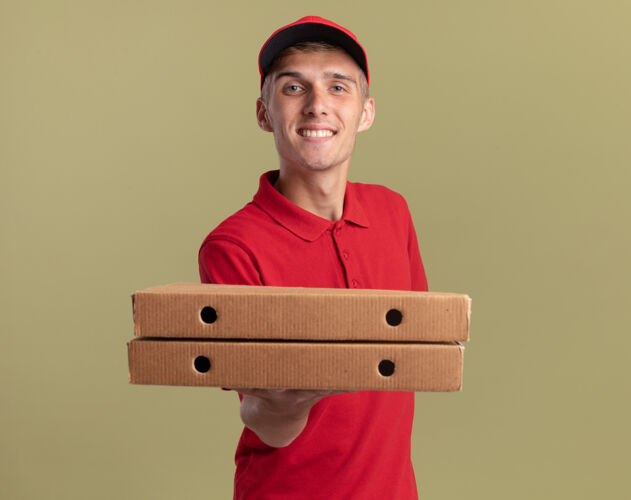 微笑微笑的年轻金发送货男孩拿着比萨饼盒看着相机年轻绿色拿着