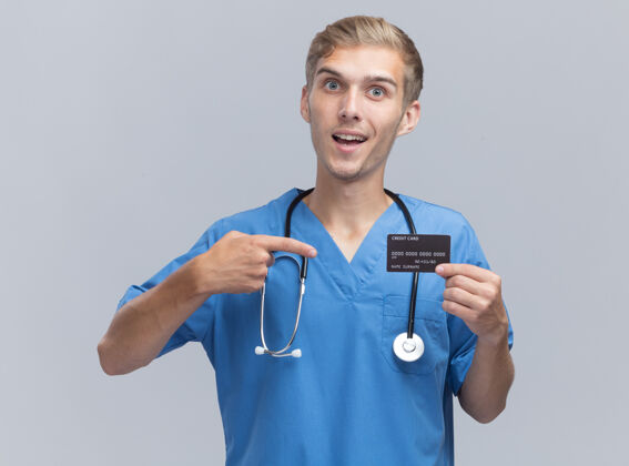 持有微笑的年轻男医生穿着医生制服手持听诊器指着隔离在白墙上的信用卡姿势穿着衣服