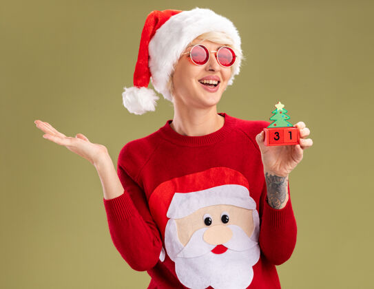 圣诞老人快乐的金发女郎戴着圣诞帽 戴着圣诞老人的圣诞毛衣 戴着圣诞眼镜 手里拿着圣诞树玩具 看起来像是在橄榄绿的墙上孤立着一只空手圣诞老人橄榄年轻