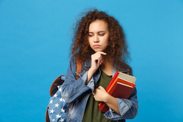 站立穿着牛仔服 背着书包的悲伤的年轻学生把书孤立地放在蓝色的墙上青年女人牛仔裤