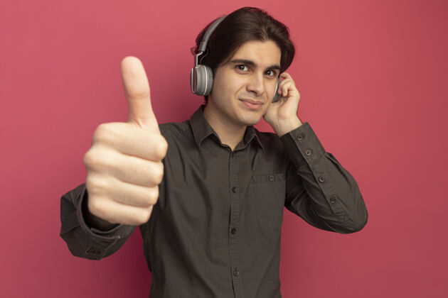 男人高兴的年轻帅哥穿着黑色t恤和耳机显示拇指向上孤立的粉红色墙上年轻表演感觉