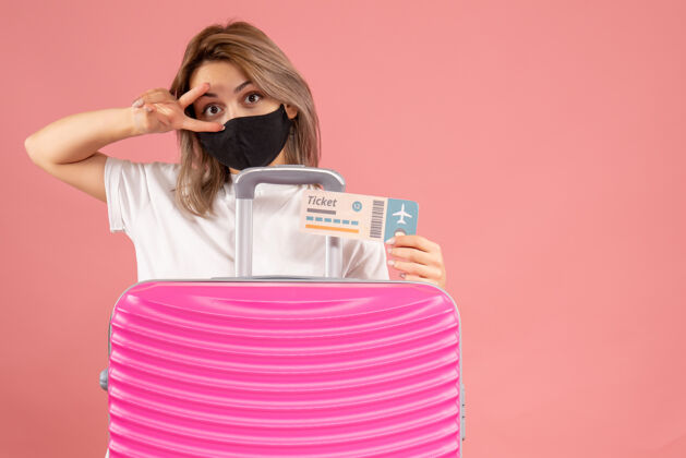 女人正面图：戴着黑色面具的年轻女子拿着票站在粉色手提箱后面旅行者行李成人