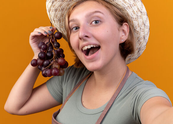 年轻高兴的年轻斯拉夫女园丁戴着园艺帽拿着葡萄看着相机自拍女葡萄采取
