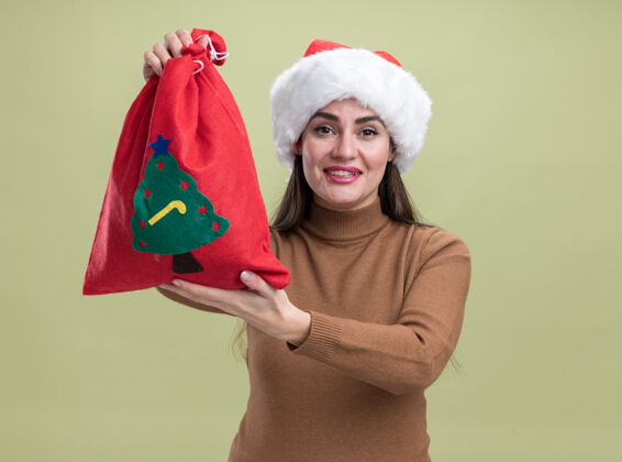 年轻微笑着的年轻漂亮女孩戴着圣诞帽 手拿着圣诞袋 孤零零地站在橄榄绿的墙上微笑抱着圣诞节