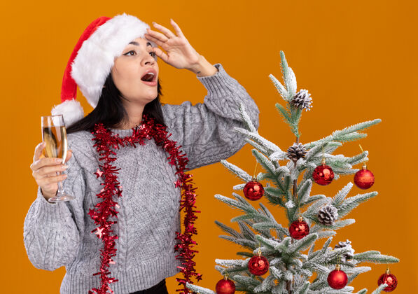 周围令人印象深刻的年轻白人女孩戴着圣诞帽 脖子上戴着金箔花环 站在装饰过的圣诞树旁 手里拿着一杯香槟 远远望着橙色墙上的隔离墙金属片年轻壁板