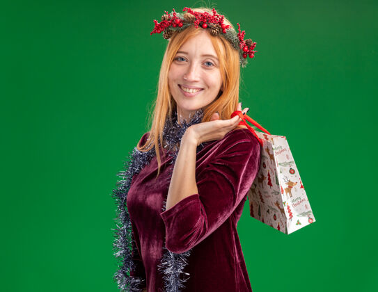 花圈微笑的年轻漂亮的女孩穿着红色的衣服 脖子上戴着花环和花环 肩上扛着礼品袋 隔离在绿色的墙上年轻圣诞节穿