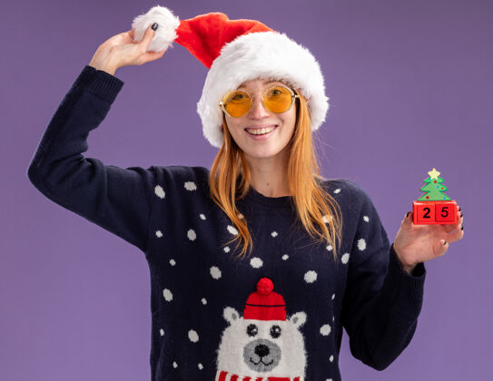 圣诞节微笑着的年轻漂亮女孩穿着圣诞毛衣 戴着圣诞帽 戴着眼镜 手里拿着圣诞玩具和帽子 隔离在紫色的墙上微笑戴着年轻人