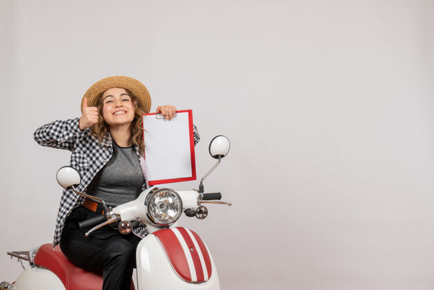 摩托车正面图：骑着轻便摩托车的年轻旅行者女孩举着红色的剪贴板竖起大拇指剪贴板人轻便摩托车