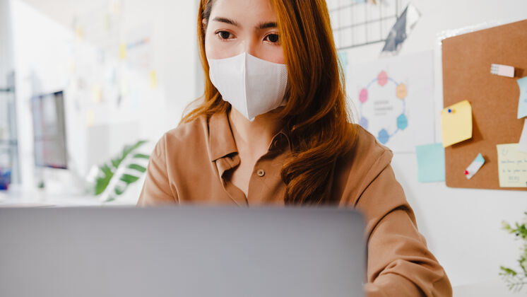 教育亚洲女商人戴口罩 在新的正常情况下保持社交距离 以防病毒 在办公室使用笔记本电脑向同事演示视频通话中的计划商务技术办公室