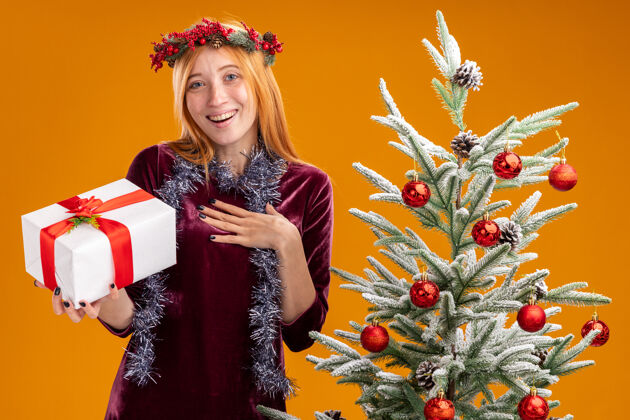 抱着微笑着站在圣诞树旁的年轻漂亮女孩 身穿红色连衣裙 脖子上戴着花环 手里拿着礼盒 手放在橙色的墙上孤立的自己身上年轻微笑衣服