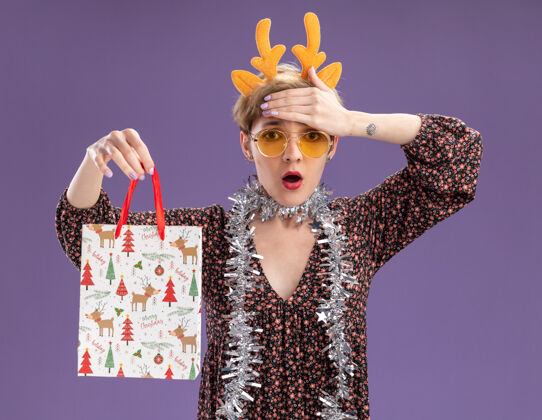 眼镜关心的年轻漂亮女孩戴着驯鹿鹿角头带 脖子上戴着金箔花环 戴着眼镜 手里拿着圣诞礼品袋 手放在额头上 隔离在紫色的墙上圣诞节漂亮周围