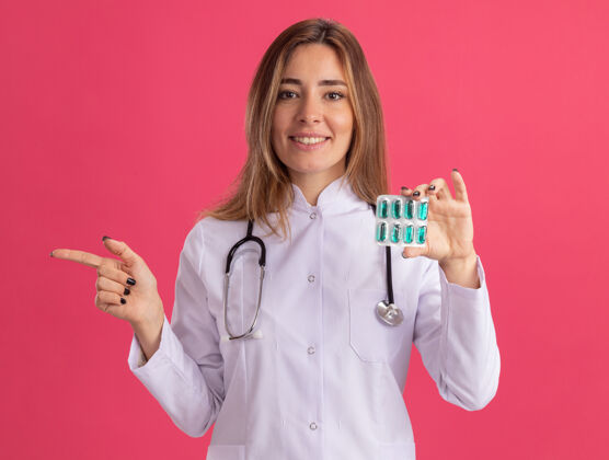 站面带微笑的年轻女医生穿着医用长袍 手持听诊器 指着隔离在粉红色墙上的药片年轻人医学人