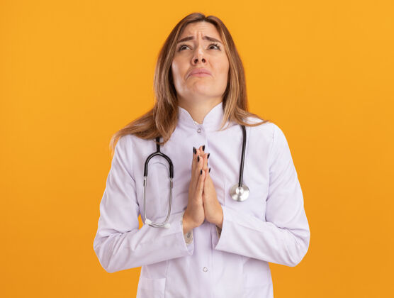 医学年轻的女医生穿着医用长袍 在黄色的墙上用听诊器做着祈祷的手势悲伤人女性