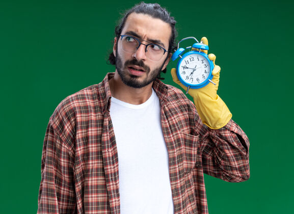 清洁年轻帅气的清洁工穿着t恤 戴着手套 拿着闹钟听绿色墙上的隔离衣服人印象