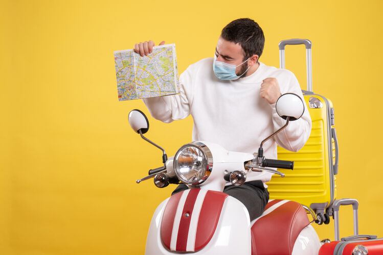 持有旅行概念的俯视图 戴着医用面罩的骄傲的家伙站在摩托车旁边 黄色的手提箱放在上面 手里拿着地图医疗球面具