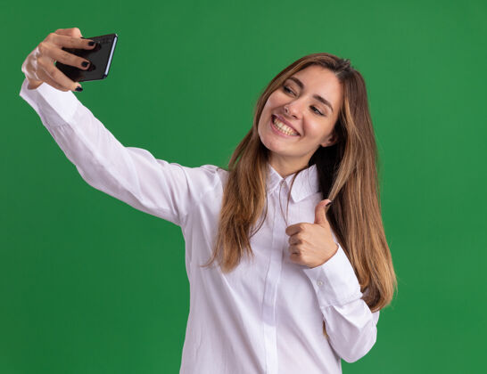 漂亮年轻漂亮的白人女孩微笑着竖起大拇指拿着手机看自拍自拍电话向上