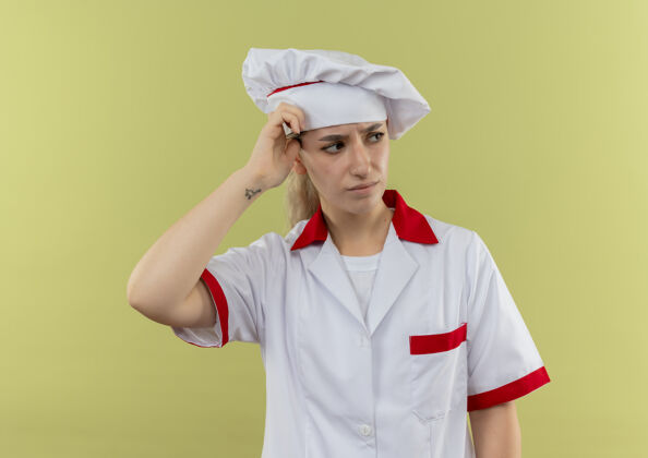 头迷茫的年轻漂亮厨师穿着厨师制服看着一边把手放在头上隔离在绿色的墙上手年轻壁板