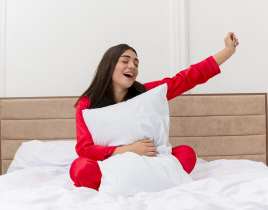 快乐穿着红色睡衣的年轻美女坐在床上 带着枕头 快乐而兴奋地在卧室里享受周末周末卧室红色