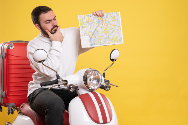 微笑顶视图自信的年轻人坐在摩托车上 手提箱上拿着地图 背景是孤立的黄色手提箱坐着摩托车