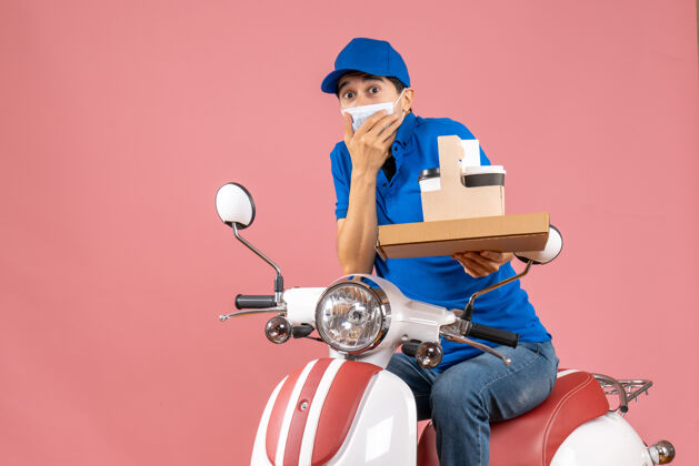 车辆困惑的男性送货员戴着口罩 戴着帽子 坐在滑板车上 在柔和的桃色背景下展示订单桃困惑摩托车
