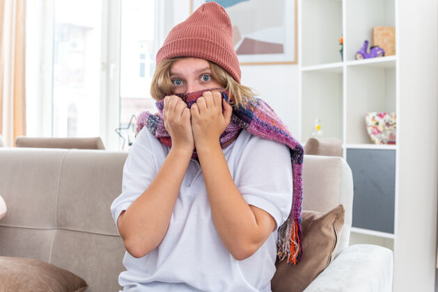 生活不健康的年轻女子戴着帽子 脖子上围着暖和的围巾 感觉不舒服 生病 患感冒和流感 坐在客厅里的沙发上 看起来很担心担心周围年轻