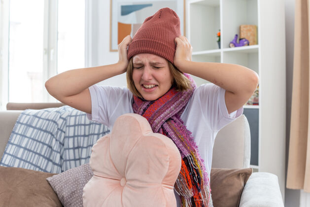 痛苦不健康的年轻女子戴着暖和的帽子 戴着围巾 看起来不舒服 摸着头 发烧 头痛 患感冒和流感 坐在明亮的客厅沙发上冷沙发流感