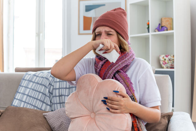 生活不健康的年轻女子戴着暖和的帽子 戴着围巾 用纸巾擤鼻涕 打喷嚏 患感冒和流感 坐在明亮的客厅椅子上鼻子帽子椅子