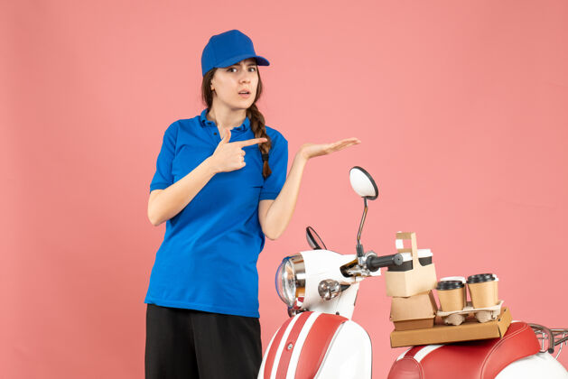 立场俯视图：快递员女士站在摩托车旁边 车上有咖啡和小蛋糕 在柔和的桃色背景上指向左边的东西壁板桃子背景