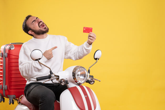 黄色旅游概念与年轻的快乐旅行男子坐在摩托车上 手提箱上指着黄色的银行卡旅游男人旅游球员