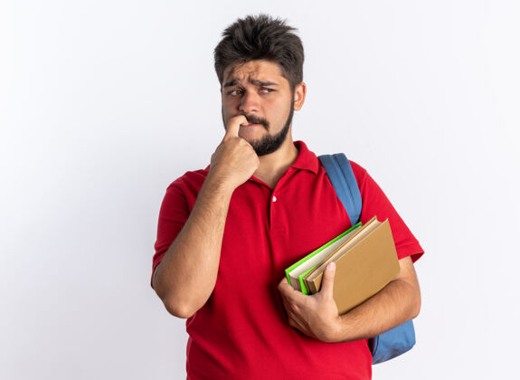 马球年轻的留着胡子的学生 穿着红色马球衫 背着书包 拿着笔记本 看着一边 紧张的咬着指甲站在白色的墙上年轻人人手势
