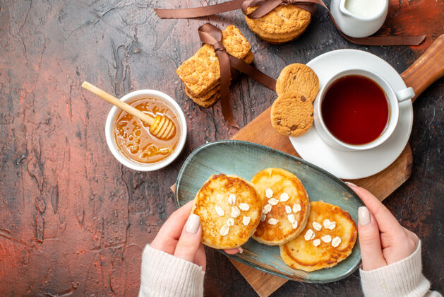 服用手拿托盘和新鲜煎饼的特写镜头一杯红茶放在木制砧板上蜂蜜饼干牛奶堆在黑暗的表面上牛奶蜂蜜晚餐