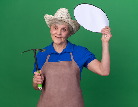 女高兴的上了年纪的女园丁戴着园艺帽拿着锄头耙和话泡帽子园艺耙子
