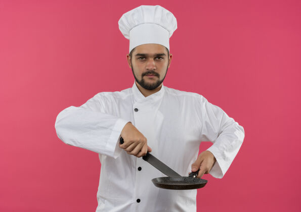 信心自信的年轻男性厨师身着厨师制服 手持刀和煎锅 隔离在粉红色的墙上烹饪平底锅年轻