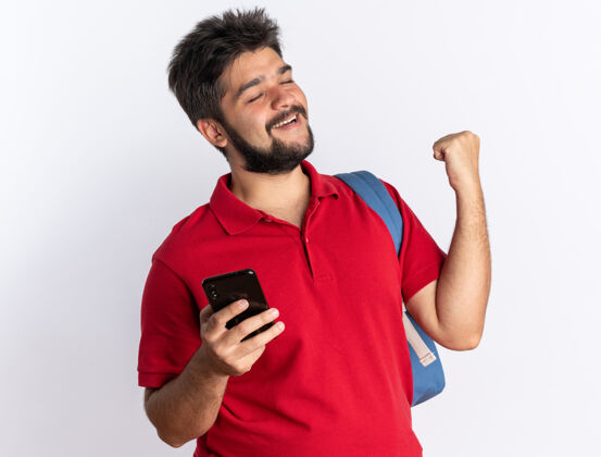 拳头年轻的留着胡子的学生 穿着红色马球衫 背着背包 手里拿着智能手机 握紧拳头 站在白色的墙上 快乐而兴奋背包兴奋人