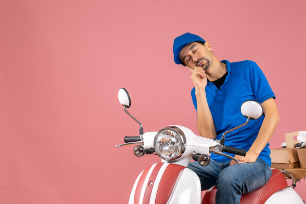 桃睡意朦胧的快递员戴着帽子坐在粉彩桃色背景上的滑板车上摩托车滑板车坐