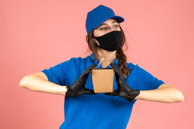 相机半身镜头忙碌的信使女孩戴着医用口罩和手套拿着小盒子在粉彩桃背景上摆姿势拍照背景成人身体