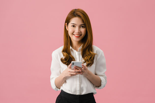 购物惊讶的亚洲小姐用手机积极的表情 笑容灿烂 穿着休闲服 看着粉色背景的相机年轻商务女性互联网